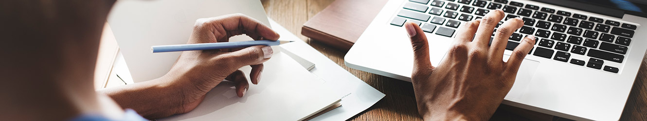 Woman writing with left hand, using computer trackpad with right hand.